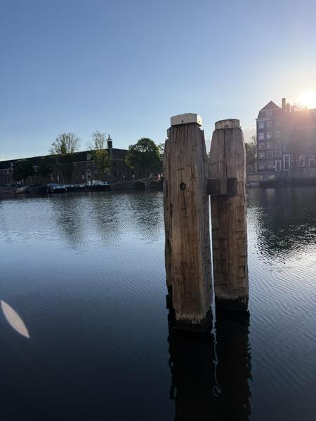 This photo captures a serene morning view of the Amstel canal in Centrum, Amsterdam, with two prominent wooden mooring posts in the foreground